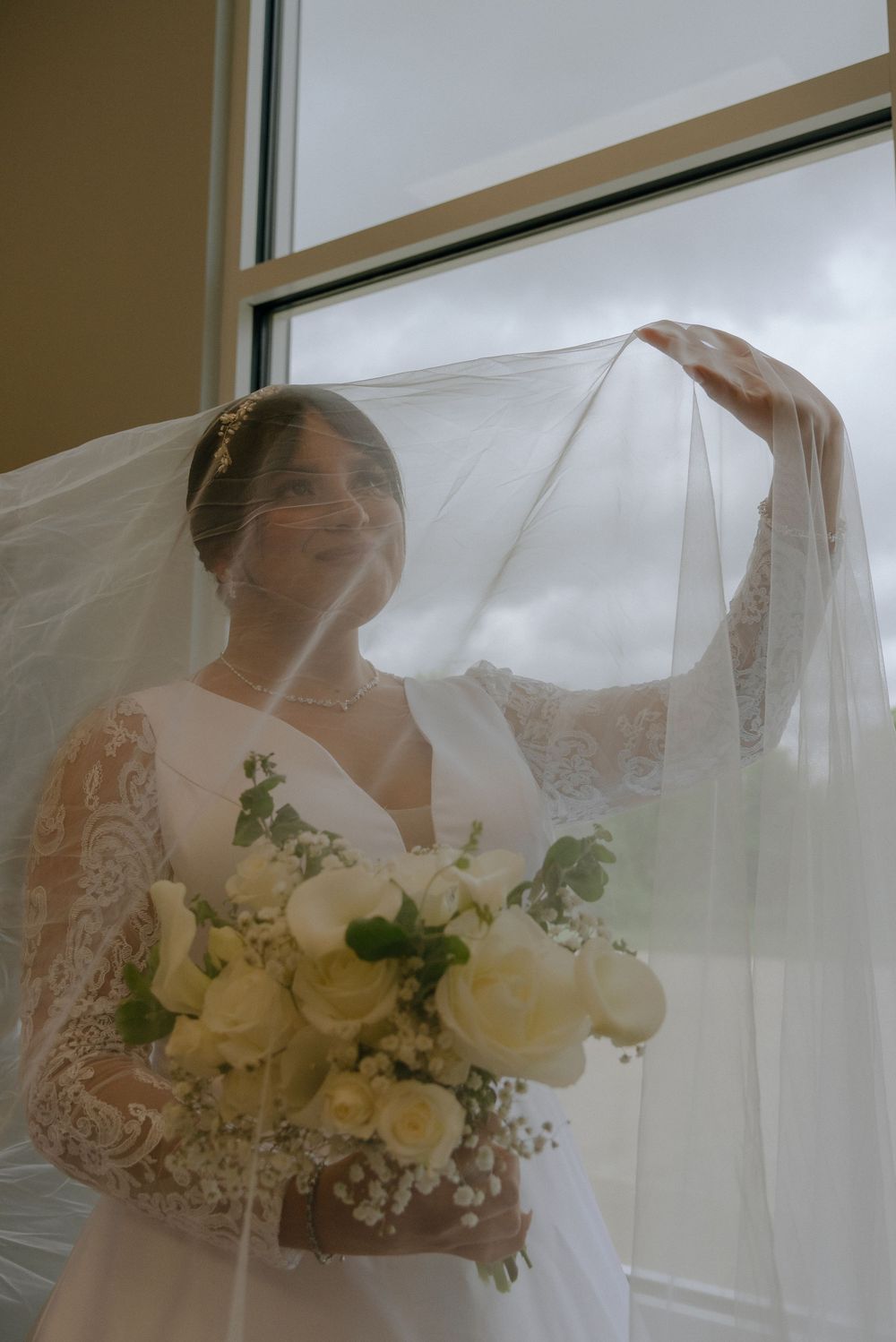 Bride in lace wedding dress holds white floral bouquet, veil flowing, standing by window on her wedding day.
