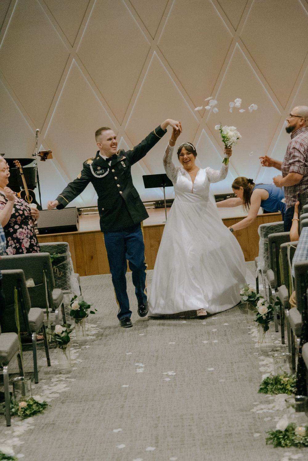 Joyful newlyweds walk down the aisle, groom in military uniform, bride in white gown holding bouquet.