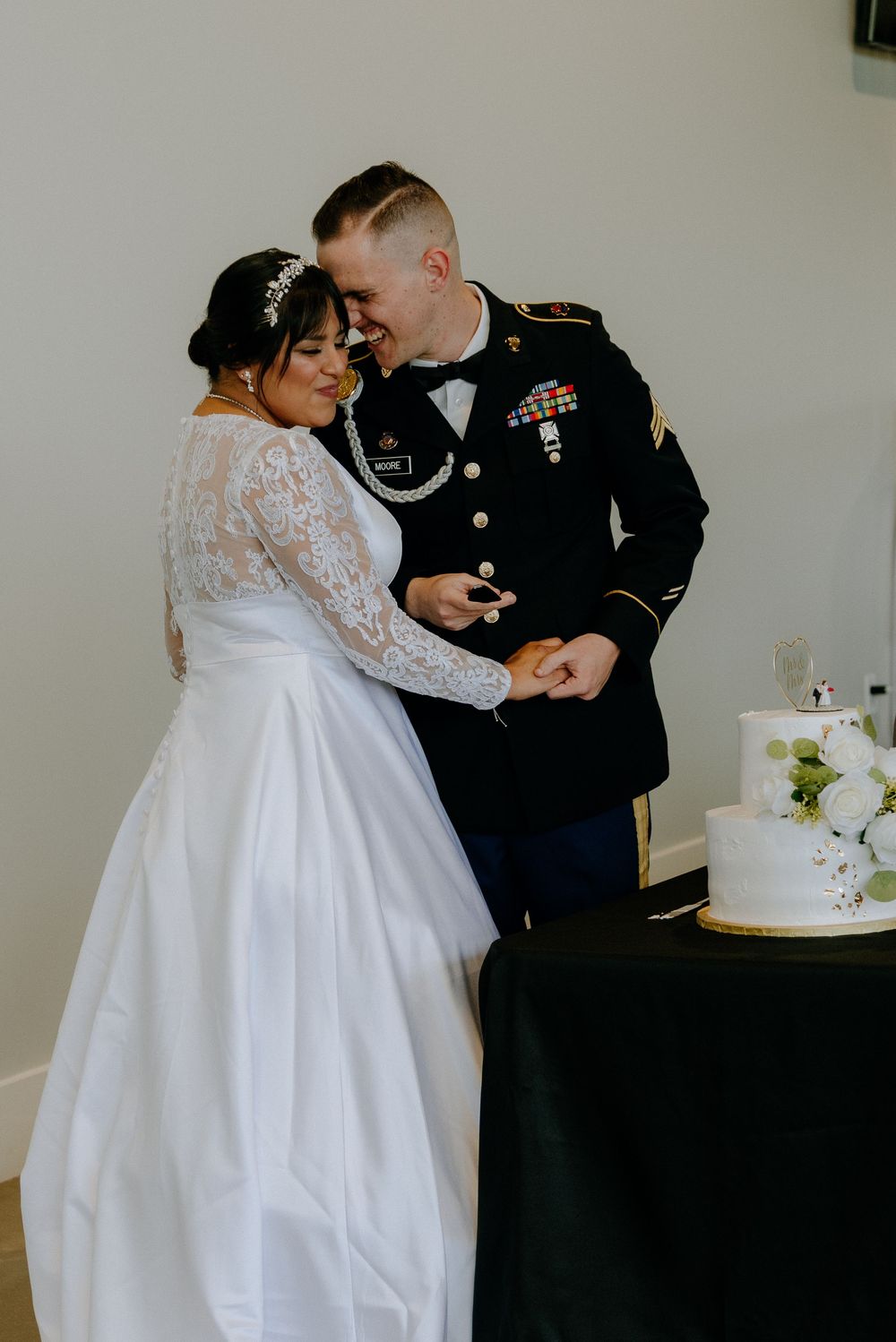 Military groom in dress uniform and bride in lace wedding gown cutting their tiered white wedding cake together.