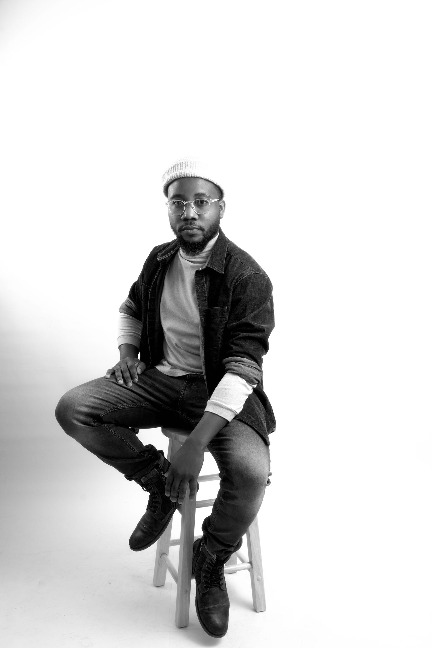 Young Black man in white shirt and black pants sits on wooden stool against yellow background.