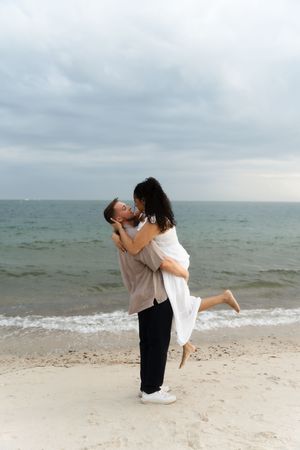 Romantic couple kissing on a sandy beach, man lifting woman in white dress,  Located in Connecticut at Harkness State Park