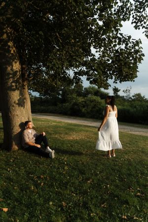 A man sits against a large tree while a woman in a white dress stands nearby in a lush green park at sunset. Flower field located in Connect
