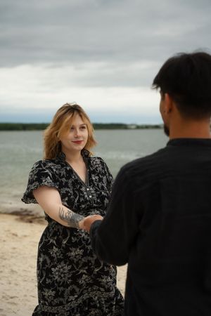 Couple holding hands on a sandy beach, woman in floral black dress with tattoo, overcast sky backdrop. McCook Beach in Niantic Connecticut