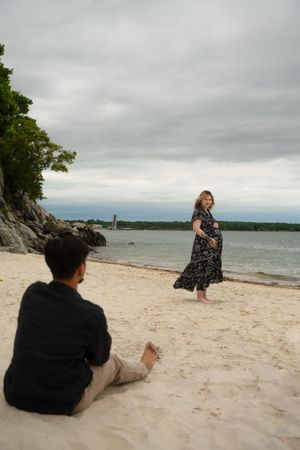 Pregnant woman in floral dress stands on sandy beach while man sits in foreground on cloudy day. McCook Beach in Niantic Connecticut