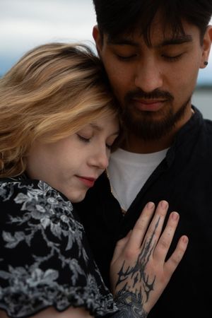 Couple embraces tenderly, woman rests head on tattooed man's chest with eyes closed on overcast day. McCook Beach in Niantic Connecticut