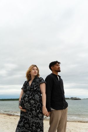 Pregnant couple holding hands on a sandy beach under cloudy skies, woman in floral dress, McCook Beach in Niantic Connecticut