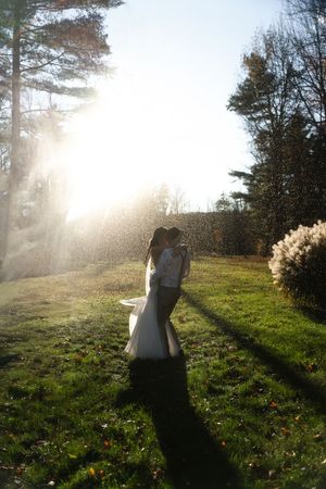 Bride and groom embracing in golden backlit sunlight on green lawn surrounded by trees. rain session in Westerly Rhode Island.