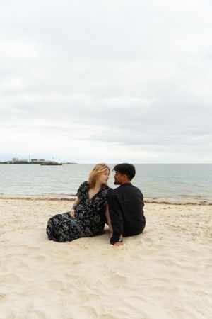 Couple sitting together on sandy beach, facing each other romantically, with calm ocean and cloudy sky McCook Beach in Niantic Connecticut