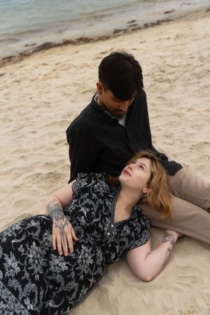 Couple on sandy beach, woman with tattoos in floral dress lying on man's lap near ocean shore. McCook Beach in Niantic Connecticut