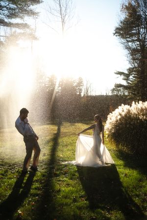 Bride in white gown and groom stand in sunlit garden with trees rain session in Westerly Rhode Island.