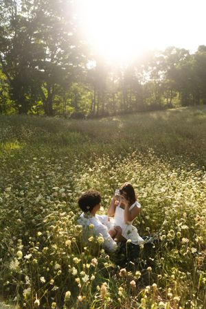Two children in white outfits sit together in a sunlit wildflower meadow surrounded by lush green trees. Flower field located in Connecticut