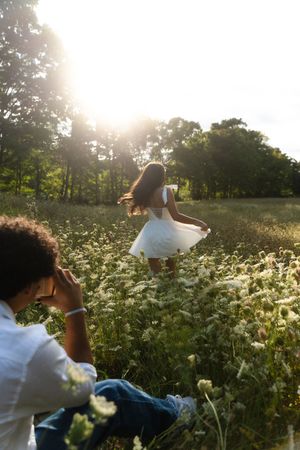 Person photographing a woman in white dress dancing in a sunlit wildflower meadow surrounded by trees. Flower field located in Connecticut