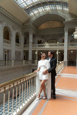 Elegant couple in wedding attire posing on a balcony inside a grand classical building with ornate architecture. Hartford city hall