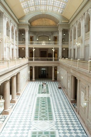 Bride and groom share a romantic moment in a grand neoclassical hall with marble floors and ornate balconies.
