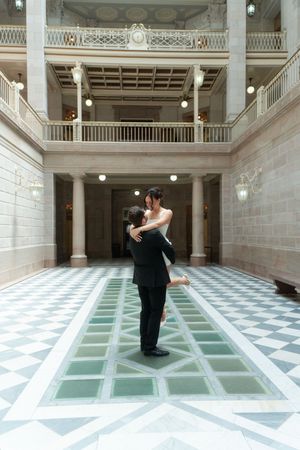 Couple embraces in elegant beaux-arts building atrium with checkered marble floors and classical columns.