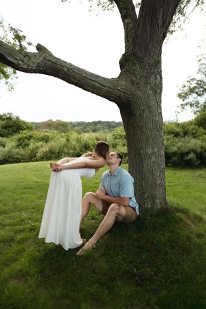 Man sitting against a tree kissing a woman in a white dress in a lush green outdoor setting.