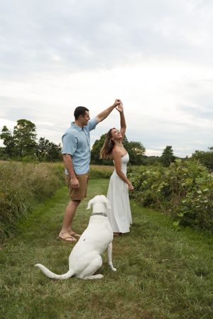 Couple dancing barefoot in a green meadow path with their white Labrador sitting nearby.