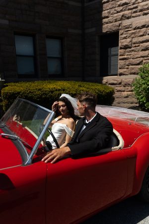 Newlywed couple in wedding attire riding in a classic red convertible sports car. Ocean Cliff in Newport Rhode Island