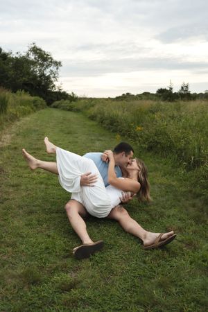 Couple sharing a kiss while lying together on a grassy path surrounded by a scenic meadow.