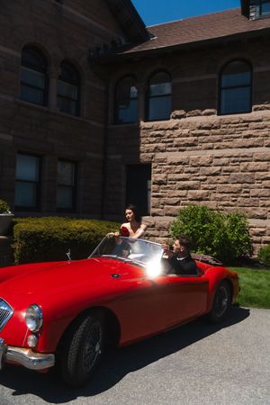 Couple posing in a classic red MGA convertible sports car outside a stone building on a sunny day. Ocean Cliff in Newport Rhode Island