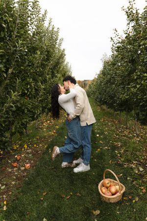 Couple kisses and embraces between rows of apple trees in an orchard, with a basket of freshly picked apples resting on the grass.
