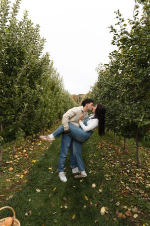 Couple kissing in an apple orchard as he dips her in his arms, surrounded by rows of trees and fallen leaves on the grass.