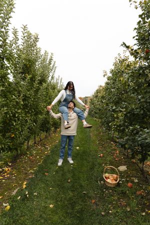 A woman rides on a man's shoulders between rows of apple trees in an orchard, with a wicker basket of apples on the grass.