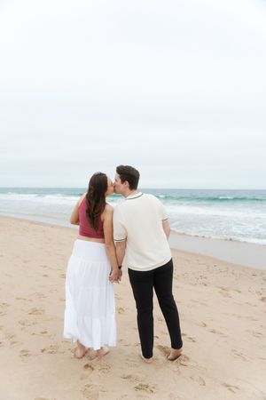 Couple holding hands and kissing on a sandy beach with ocean waves Watch Hill Beach in Rhode Island.