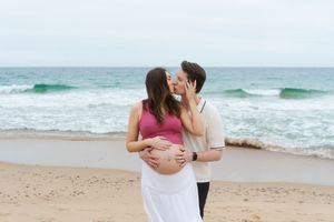 Couple kissing on beach, man embracing pregnant woman's belly, ocean waves in background. Watch Hill Beach in Rhode Island.