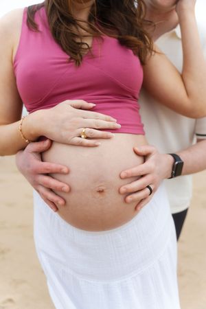 Couple cradling pregnant belly on beach, woman in pink Watch Hill Beach in Rhode Island.