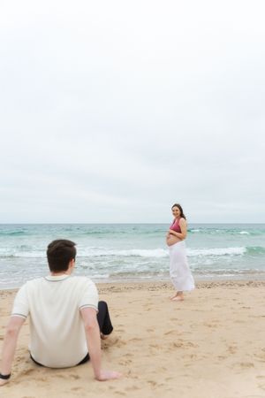 Pregnant woman in pink top and white skirt stands by ocean waves while man sits on sandy beach. Watch Hill Beach in Rhode Island.