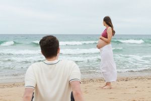 Pregnant woman in white skirt stands at ocean shore while man sits on sandy beach behind her. Watch Hill Beach in Rhode Island.