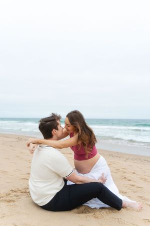 Pregnant couple sharing a kiss while sitting on sandy beach with ocean waves in the background. at Watch Hill Beach in Rhode Island.