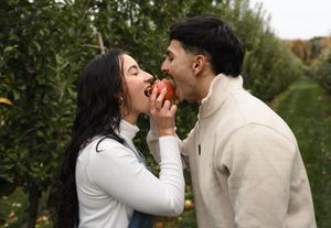 Couple in an apple orchard sharing a bite of a red apple while surrounded by green trees and rows of fruit.