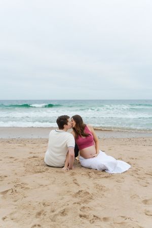 Pregnant couple sharing a kiss while sitting on sandy beach at Watch Hill Beach in Rhode Island