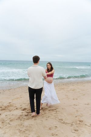 Pregnant woman in white skirt and red top holds hands with man at Watch Hill Beach in Rhode Island.