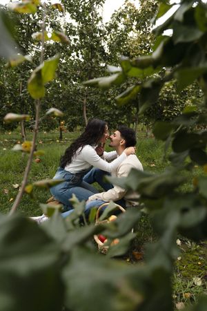 Couple sharing an intimate moment while seated on the grass in a sunny apple orchard, framed by green leaves in the foreground.