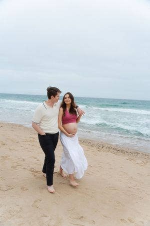 Couple walking on sandy beach, pregnant woman in pink at Watch Hill Beach in Rhode Island.