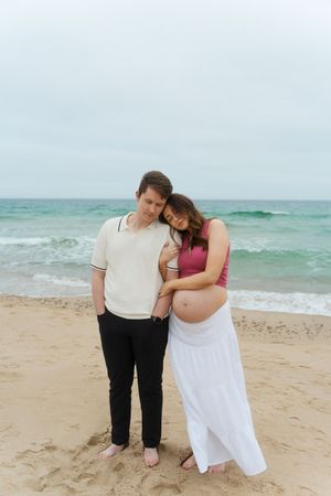 Pregnant woman in pink crop top leans on partner's shoulder at Watch Hill Beach in Rhode Island.