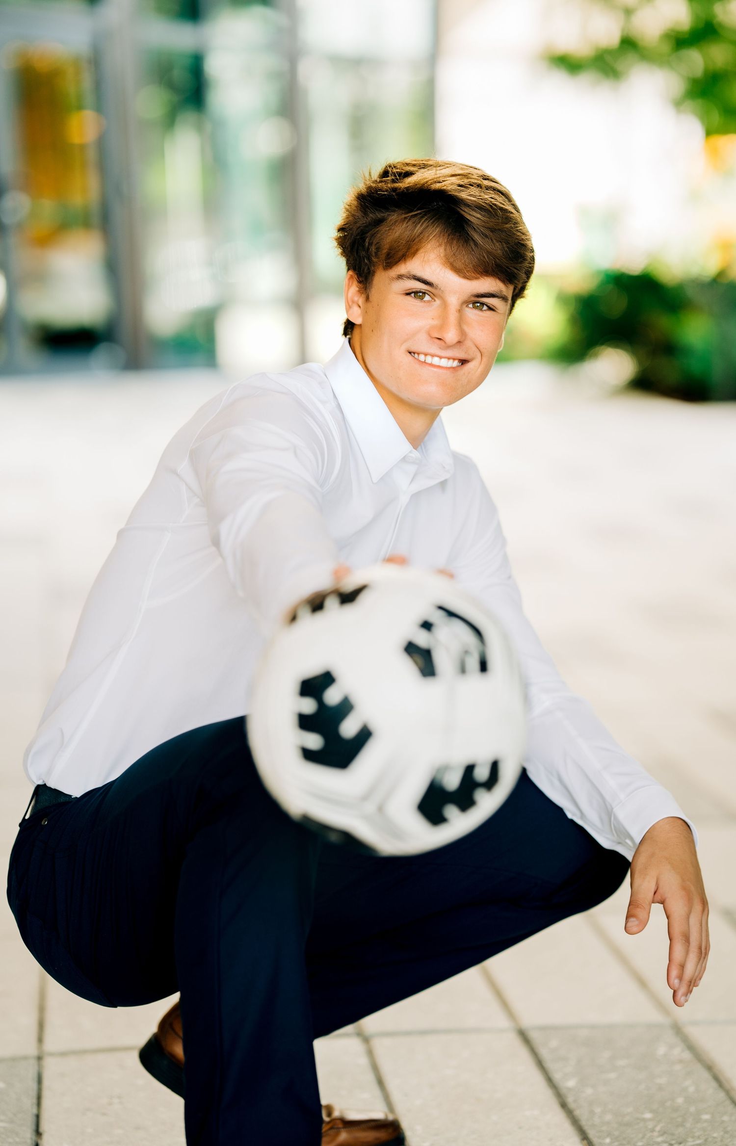Smiling young man in white shirt crouching outdoors, balancing a soccer ball on his knee for senior photos.