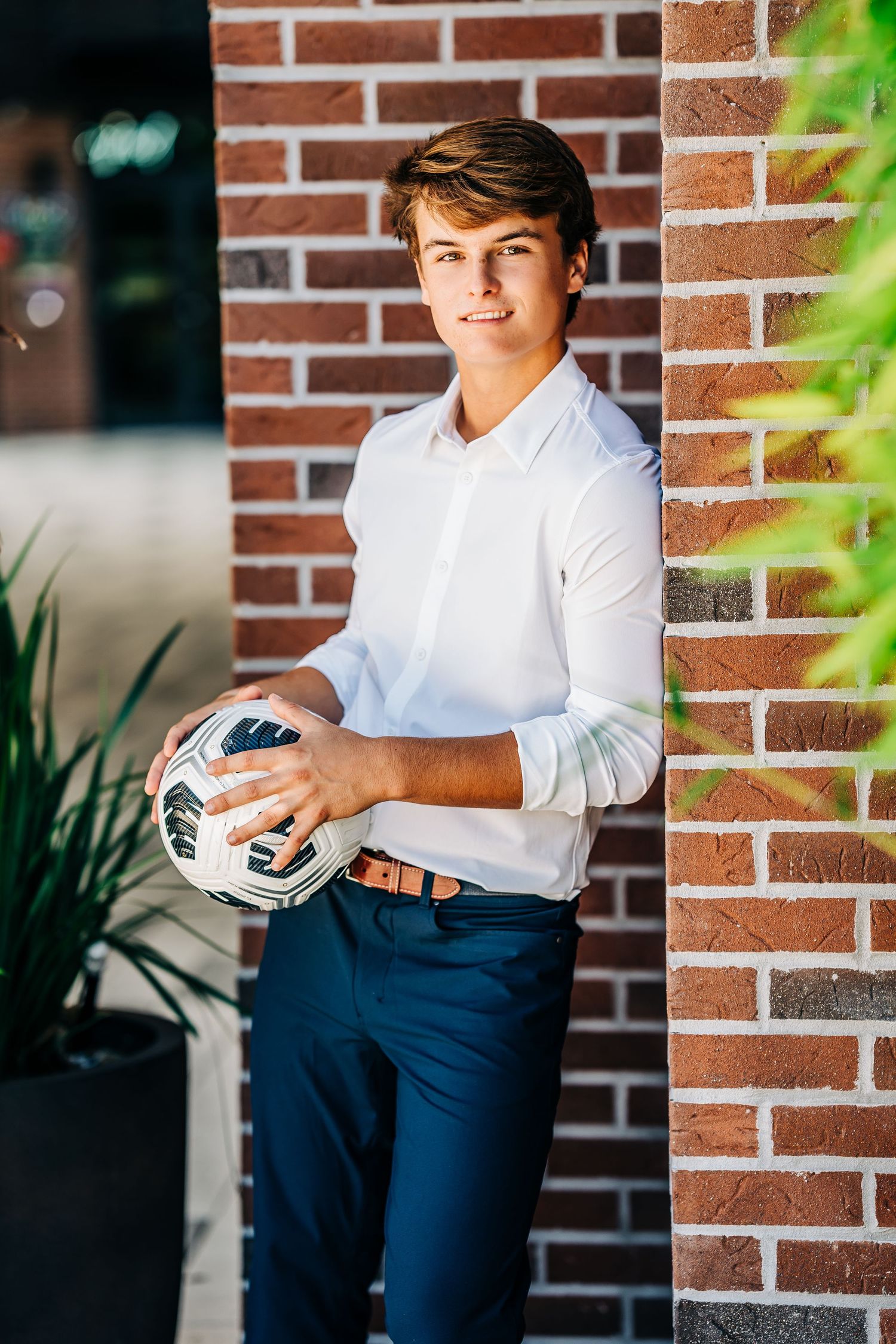Young man in white shirt holding soccer ball leaning against brick wall outdoors.