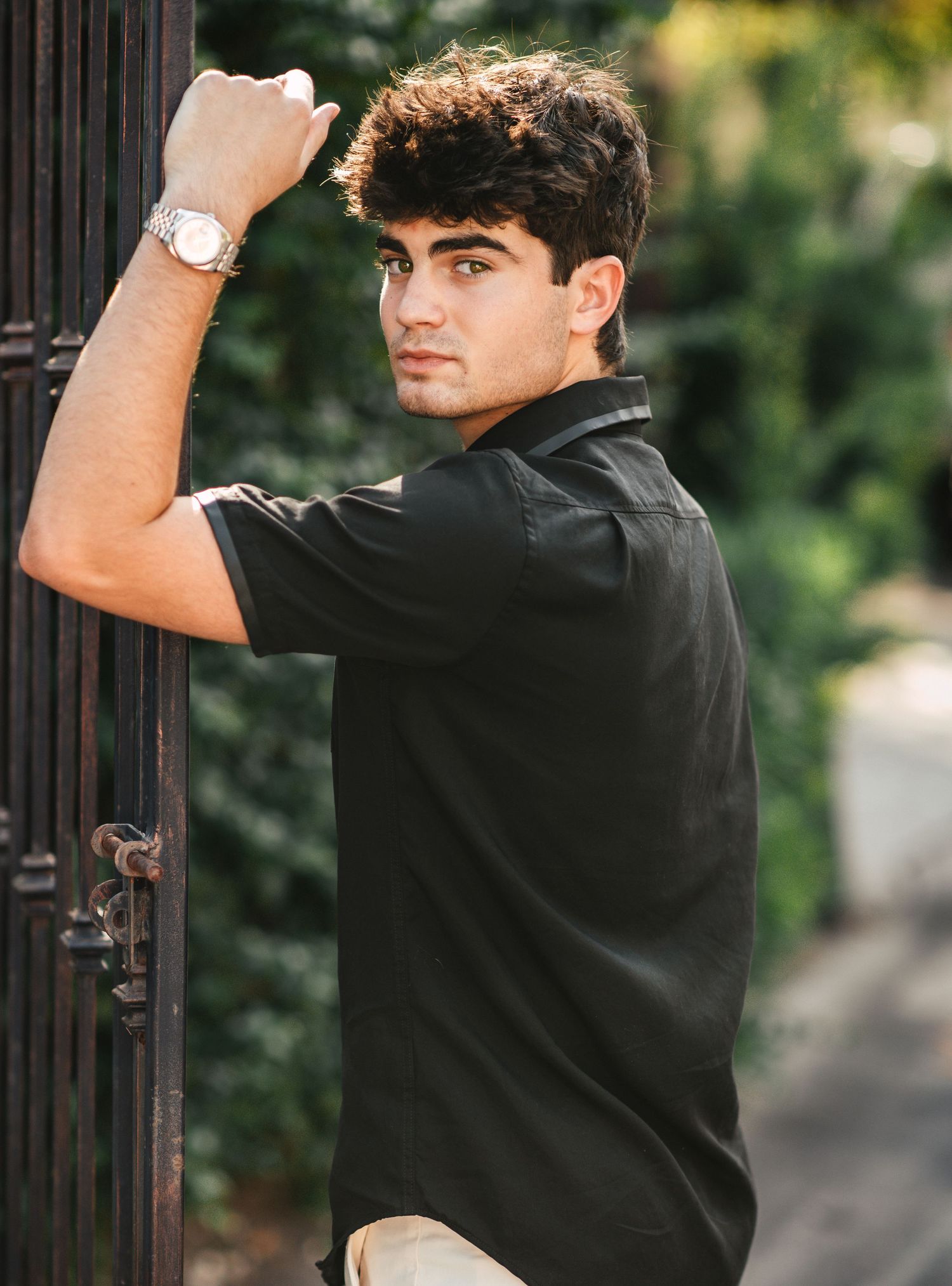 Young man in black shirt and silver watch leans against iron gate, posing outdoors with lush greenery background.