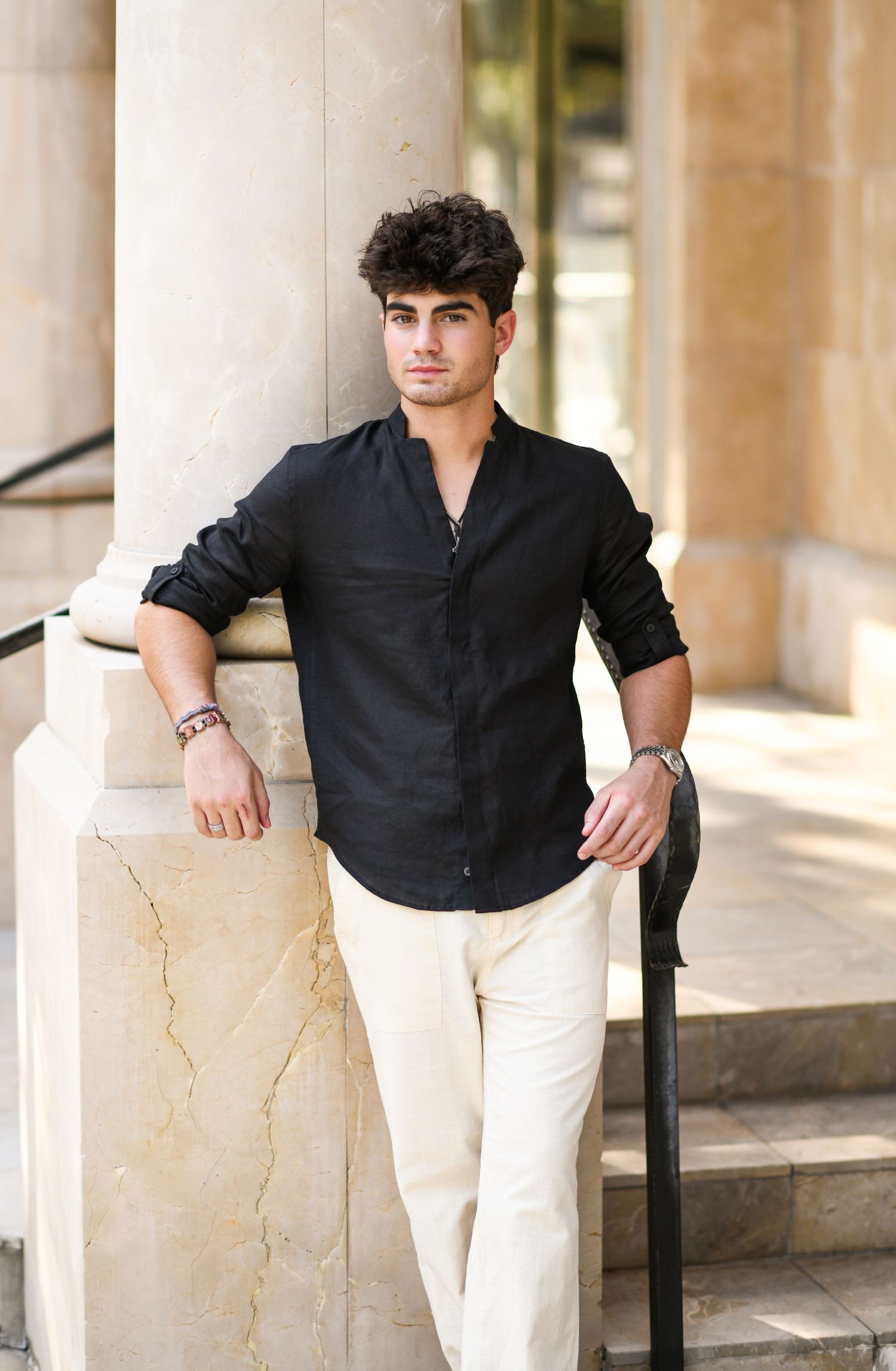 Young man in black linen shirt and white pants leaning against marble column outdoors.