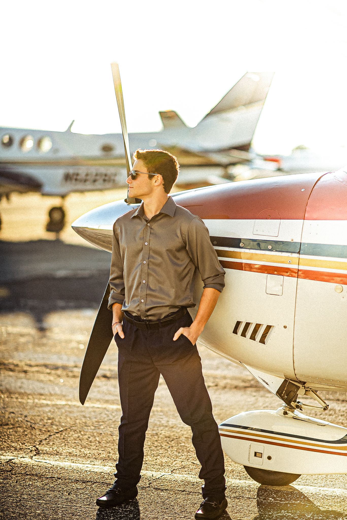 Young man in sunglasses and gray shirt stands confidently beside a small propeller aircraft on a sunny airport tarmac.