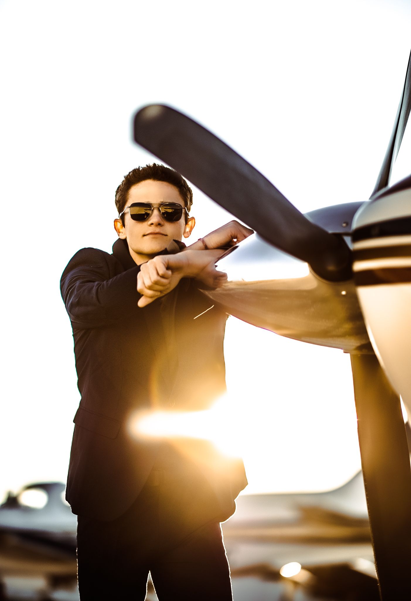 Young aviator in sunglasses and black jacket leaning on aircraft propeller with golden backlight at airfield.