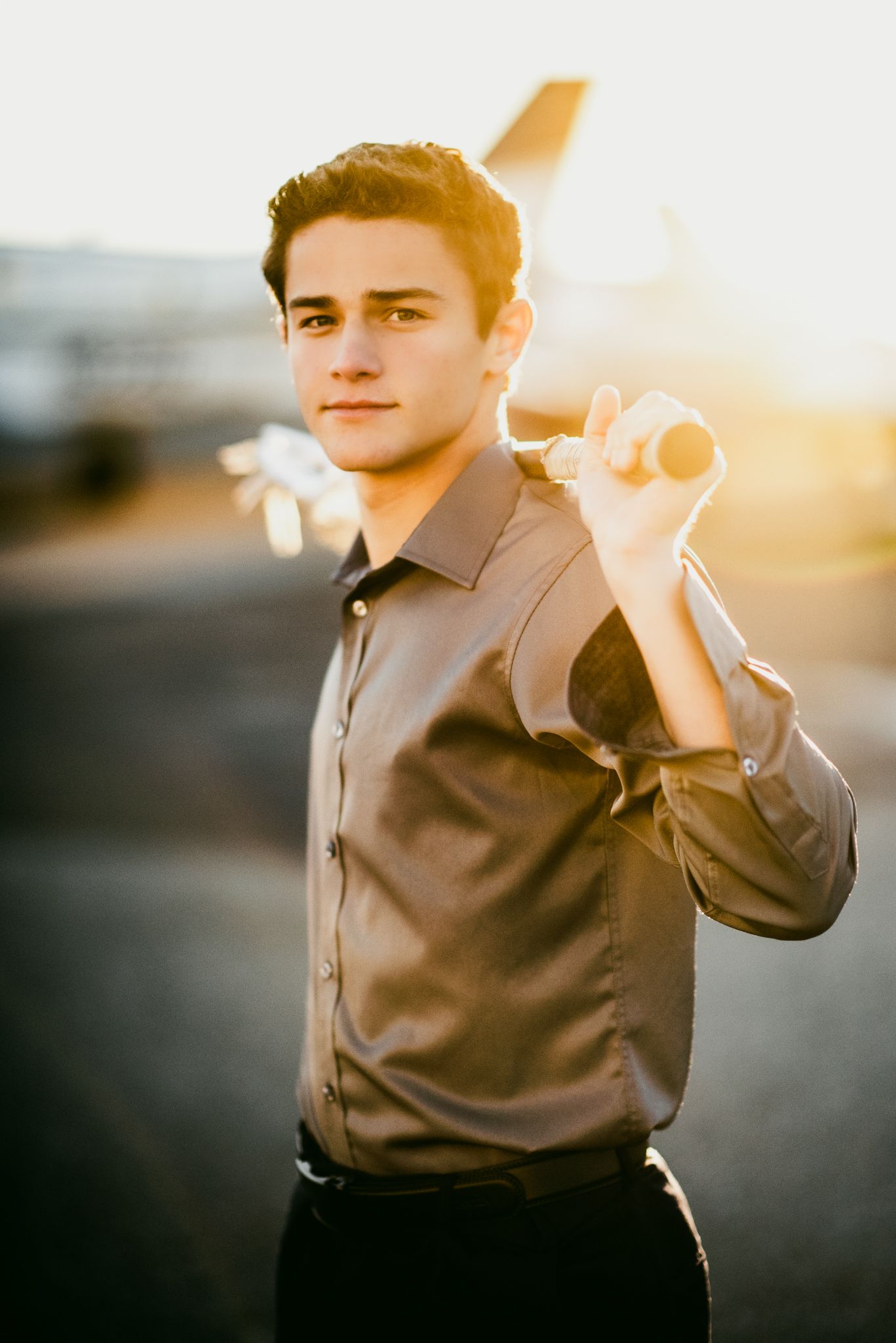 Young man in gray shirt holds model airplane at golden hour on airport tarmac with aircraft in background.