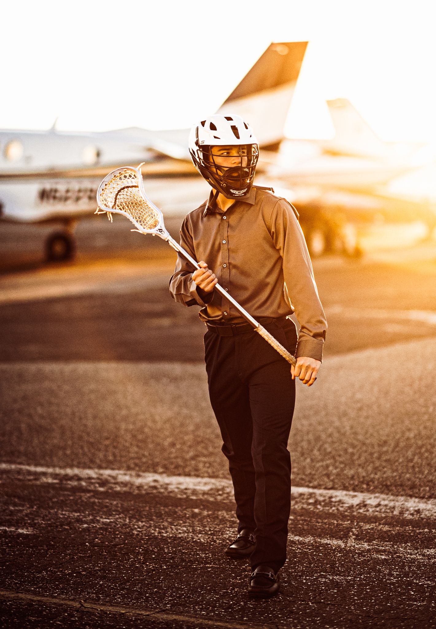 Young lacrosse player in helmet holds stick on airport tarmac with private jet at golden hour sunset.