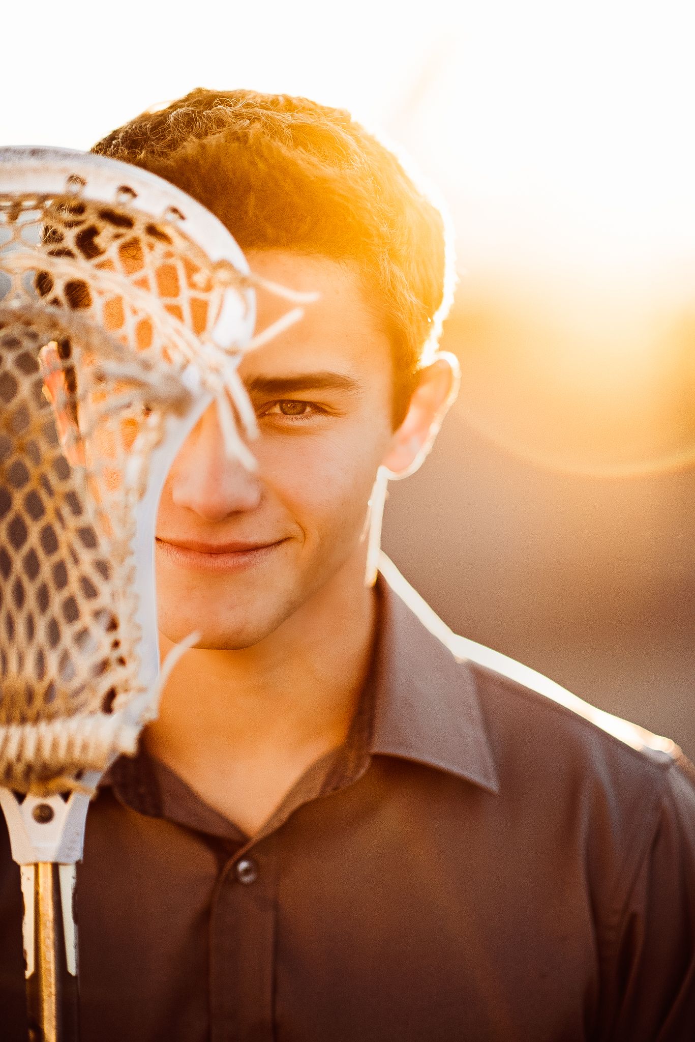 Young man holding a lacrosse stick in front of his face at golden hour, smiling softly in warm sunlight.