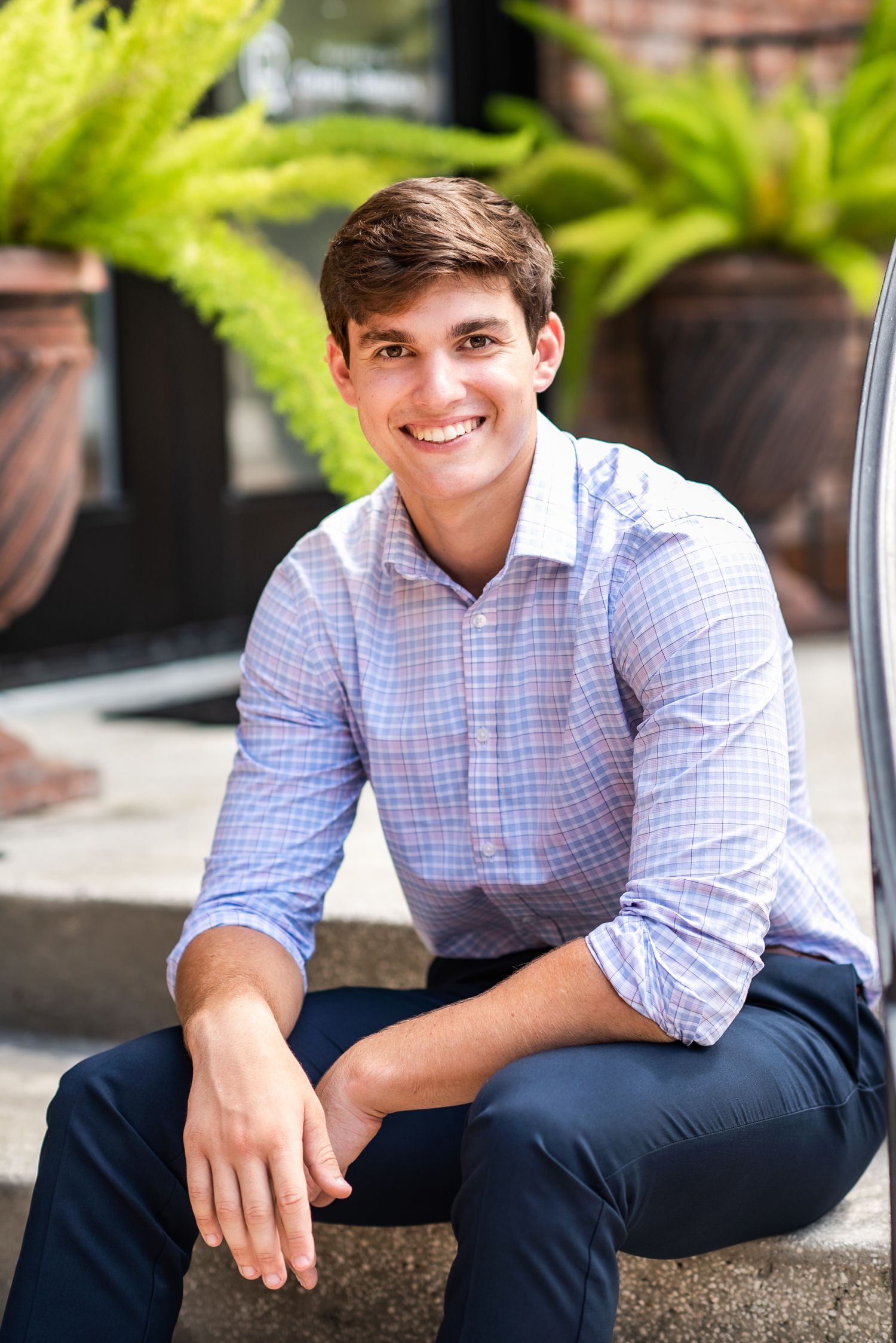 Smiling young man in plaid shirt sitting outdoors near green ferns and planters.