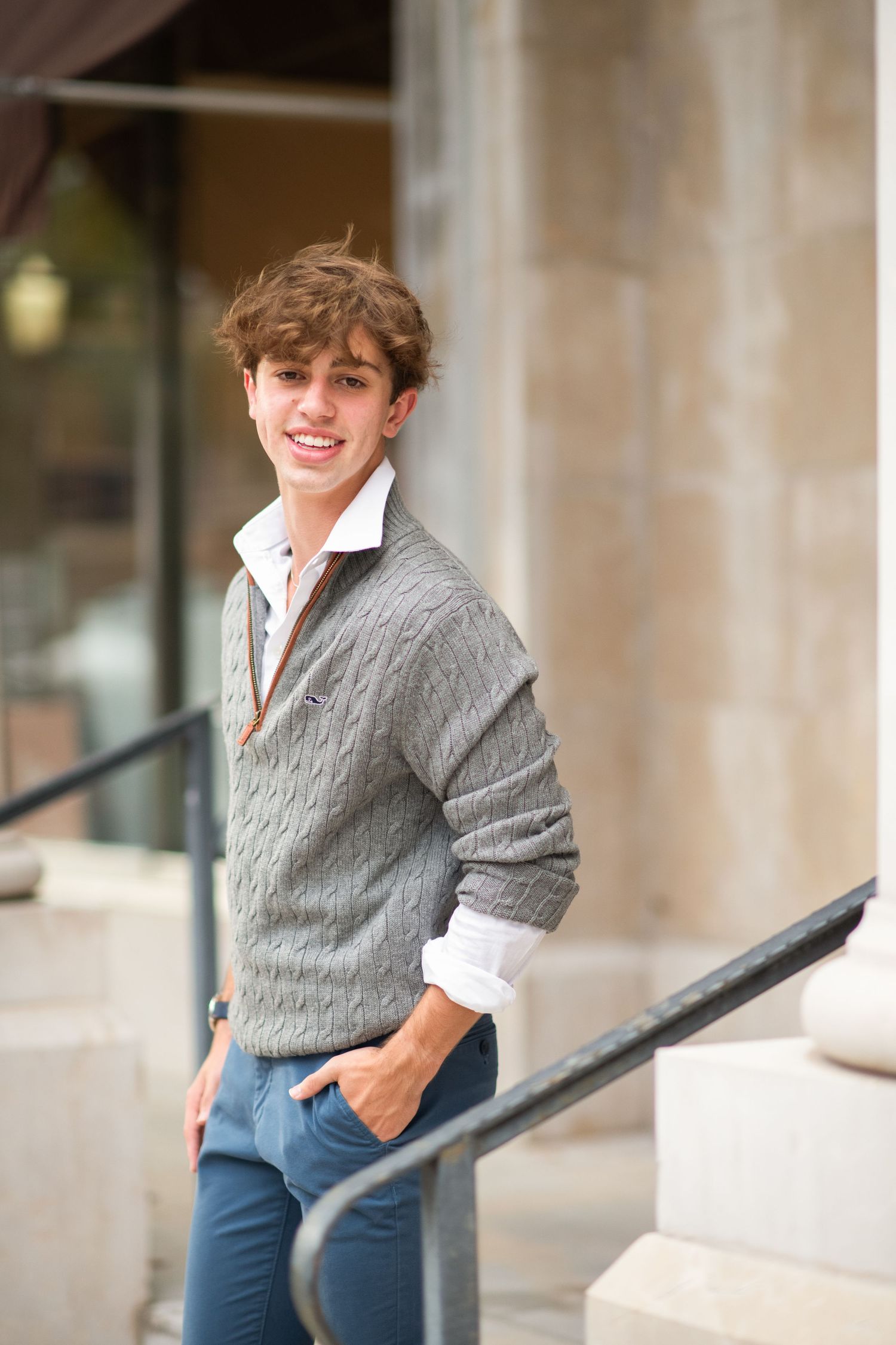 Smiling young man in gray cable-knit sweater and jeans poses on outdoor steps near a metal railing.
